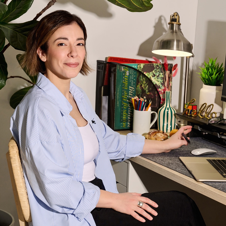 Une femme est assise à un bureau avec un ordinateur portable, une lampe, des livres, des crayons, une tasse et des pâtisseries. Elle est souriante et porte une chemise à rayures bleu clair sur un haut blanc. Il y a des plantes et une décoration "hello" sur le bureau.