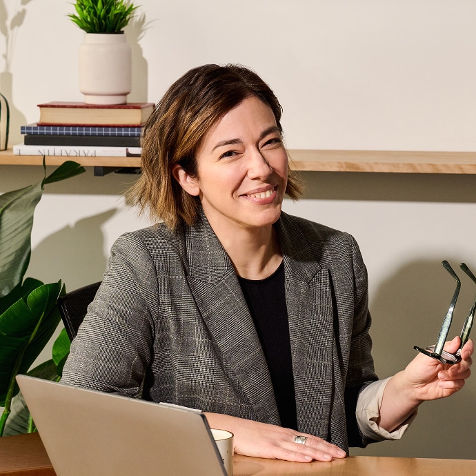 Une personne aux cheveux bruns courts, vêtue d'un blazer à carreaux, sourit en s'asseyant à un bureau et en tenant des lunettes. Un ordinateur portable, une plante, des livres et une tasse à café sont visibles sur le bureau et l'étagère derrière eux.