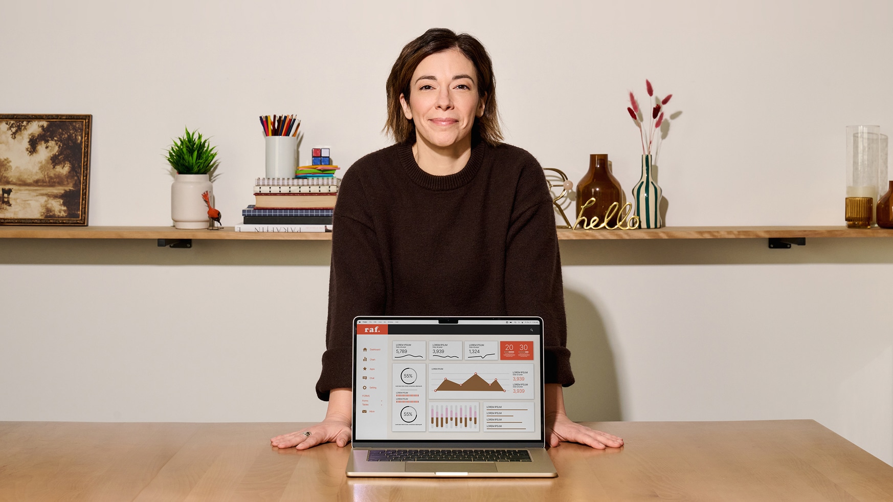 Une personne aux cheveux bruns courts et au pull marron se tient derrière une table, souriante, les mains posées sur la table. Un ordinateur portable ouvert affichant des graphiques se trouve devant eux. Une étagère avec des plantes, des livres et des objets de décoration se trouve à l'arrière-plan.