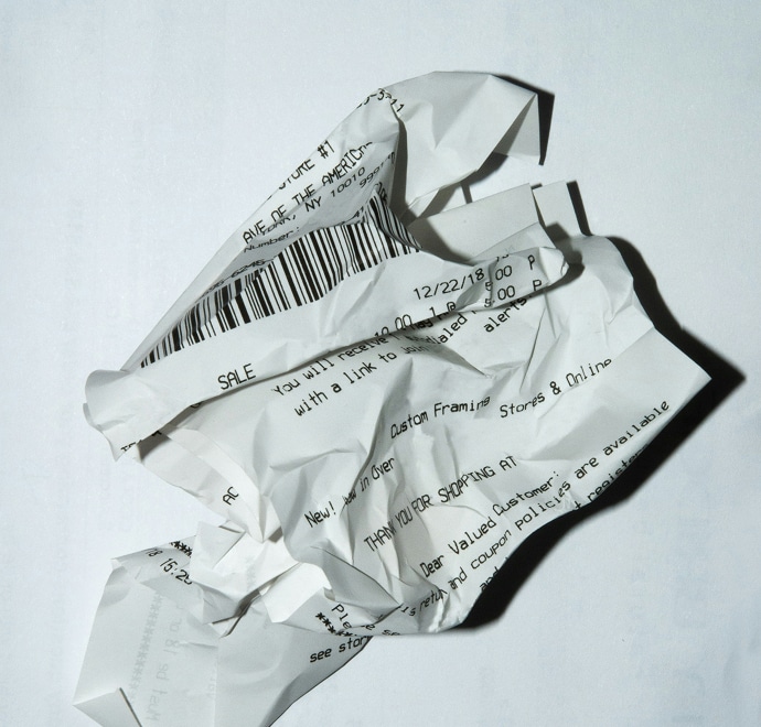 Un reçu en papier froissé avec un texte imprimé et un code-barres repose sur une surface blanche, projetant une ombre en dessous. Le texte et les détails du reçu sont partiellement visibles et déformés par les plis.