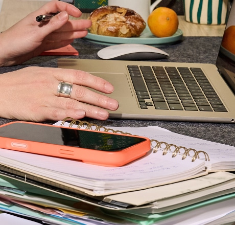 Les mains d'une personne utilisent un ordinateur portable sur un bureau encombré, avec des cahiers, un smartphone, un stylo, une pâtisserie et des fruits à proximité. La personne porte une grosse bague en argent à un doigt.