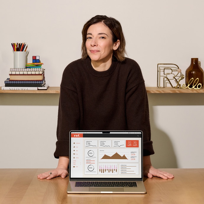 Une femme vêtue d'un pull marron se tient derrière un bureau sur lequel se trouve un ordinateur portable affichant des tableaux et des graphiques. Sur le bureau se trouvent des livres, des stylos et des objets décoratifs, dont un panneau "hello" doré et un vase marron.