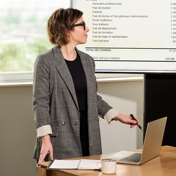Une personne portant des lunettes et un blazer à carreaux se tient à un bureau avec un ordinateur portable et un carnet de notes, et fait des gestes avec un stylo. Une grande diapositive de présentation avec un texte financier en français est affichée en arrière-plan.