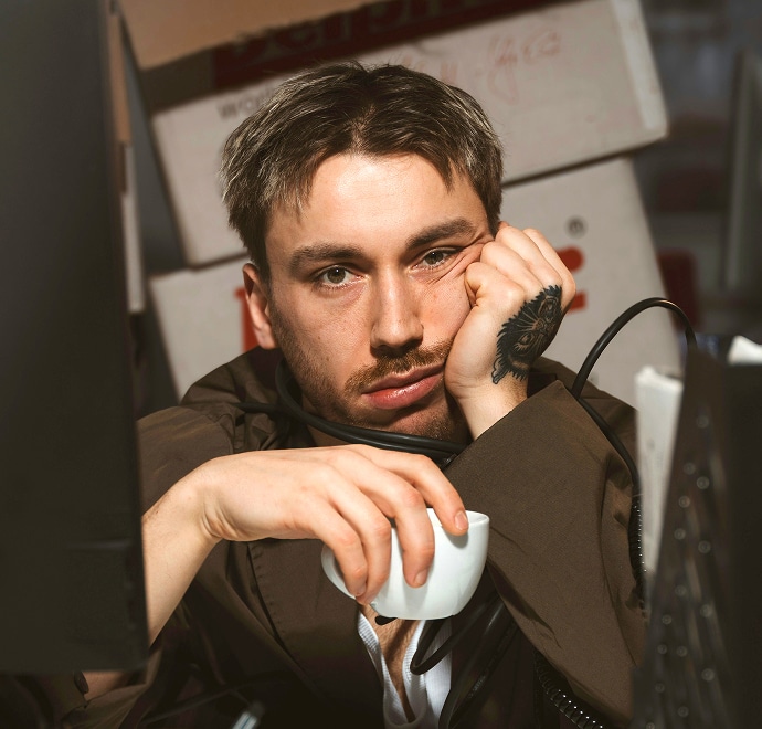 Un homme aux cheveux courts appuie son visage sur sa main, l'air fatigué ou ennuyé, tout en tenant une tasse blanche. Il est assis entre des écrans d'ordinateur, avec des boîtes en carton empilées à l'arrière-plan.