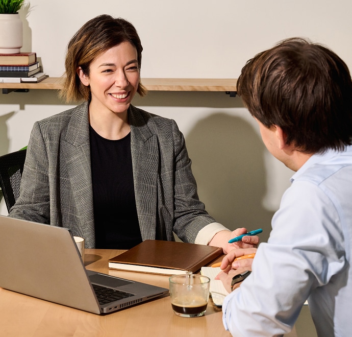 Deux personnes assises à un bureau, en train de discuter. L'une des personnes, souriante, porte un blazer gris et tient un carnet de notes. L'autre est tourné vers l'extérieur et tient un stylo. Un ordinateur portable, du café et des livres sont posés sur le bureau.