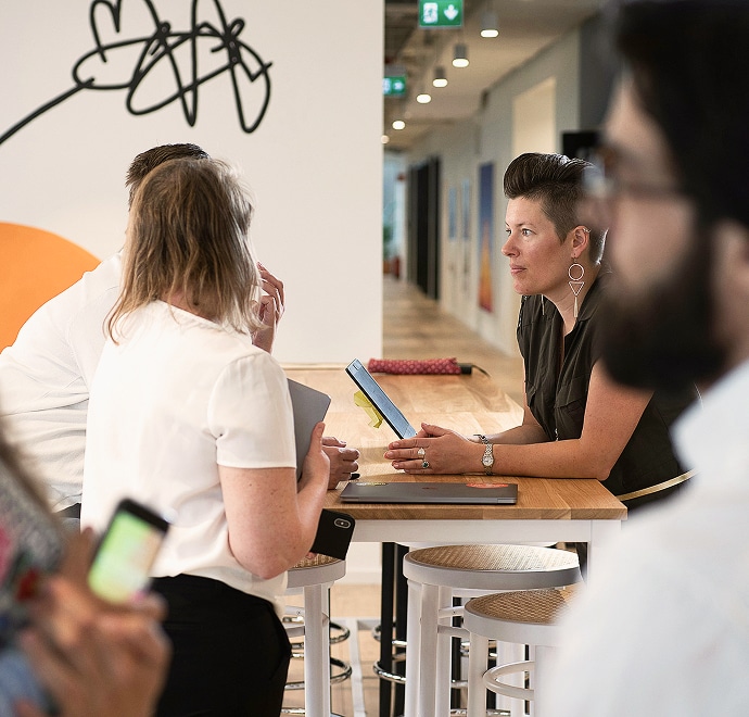 Un groupe de personnes discute autour d'une table haute dans un bureau moderne et ouvert. Une femme aux cheveux courts écoute attentivement en tenant une tablette, et d'autres personnes se tiennent à proximité, certaines tenant un téléphone.